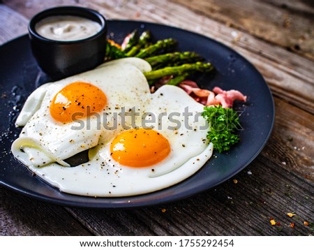 Tasty breakfast. Sunny side up eggs with green asparagus, fried bacon and garlic dip served on black plate on wooden table