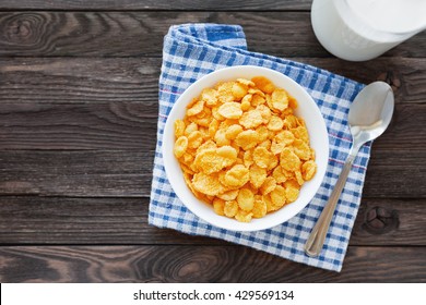 Tasty corn flakes in bowl with bottle of milk. Rustic wooden background with plaid tartan napkin. Healthy crispy breakfast snack. Place for text. Top view, flat lay.