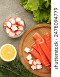 Tasty crab sticks, pieces of lemon and salad on wooden plate on grey background. Top view