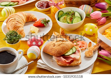 Tasty Easter brunch or spring breakfast with a fresh croissant, ham or bacon roll and coffee on a table decorated with colorful Easter eggs and tulips