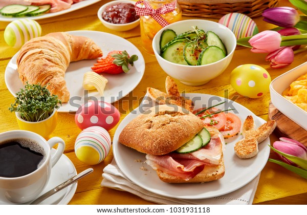 Tasty Easter brunch or spring breakfast\
with a fresh croissant, ham or bacon roll and coffee on a table\
decorated with colorful Easter eggs and\
tulips