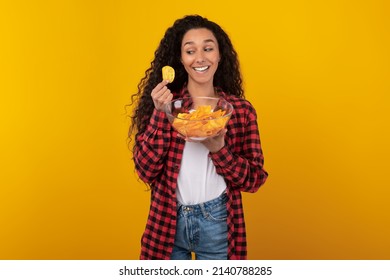 Tasty Fast Food Concept. Portrait Of Smiling Young Lady Enjoying Delicious Potato Crisps Holding Glass Bowl, Posing Looking At Chips In Hand Standing Isolated On Yellow Orange Studio Background Wall