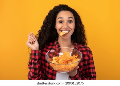 Tasty Fast Food. Portrait Of Hungry Excited Fit Female Model Eating Delicious Potato Crisps Holding Glass Bowl, Posing With Chips In Mouth Isolated Over Yellow Orange Studio Wall. Junk Meal Addiction