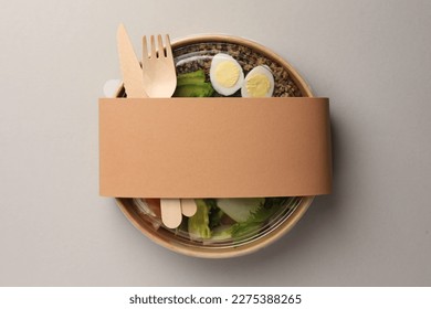 Tasty food in container with wooden fork and knife on light background, top view. Space for text