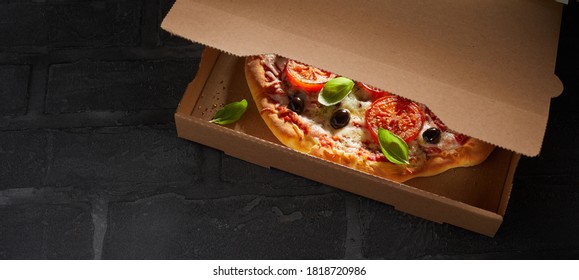 Tasty fresh oven-fired Italian pizza in a brown cardboard takeaway box with the lid partially open to reveal the lit interior on a black tiled counter with copyspace - Powered by Shutterstock