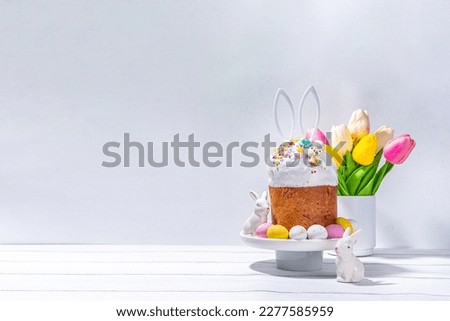Tasty Glazed Easter cake with sugar decor, colorful eggs basket, ceramic rabbits and spring flower on white kitchen table, copy space. Happy Easter Holidays greeting card background copy space