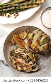 Tasty grilled artichokes served with sauce and asparagus on white wooden table, flat lay - Shutterstock ID 2386456233