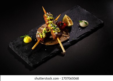 Tasty Grilled Beef Cube Steak In A Stick With Grilled Lime And Whole Tomato On The Side. Elegant Main Course Plating In Plain Black Background. Food Photography For Fine Dine Restaurant Menu.