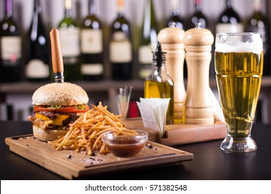 Tasty grilled cheeseburger with a knife stabbed trough on a wooden plate. Burger with pickles, cheese, tomatoes and french fries on the side. Napkins and beer on the side. Olive oil in the background.