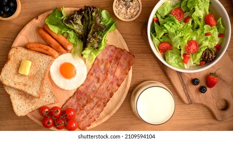 A Tasty Homemade Breakfast Served With Salad Bowl, Milk, Egg, Ham, Sausages And Toasts On Wooden Dining Table. Overhead, Flat Lay