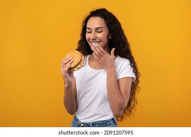 Tasty Meal. Portrait Of Happy Excited Lady Eating Big Juicy Burger, Laughing And Wiping Lips With Finger. Satisfied Woman Holding Fastfood, Isolated Over Orange Yellow Background, Ad Banner