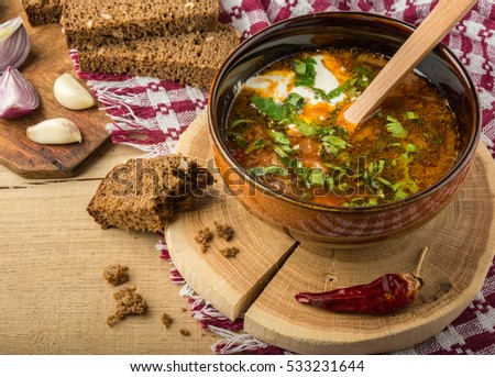 Tasty and nutritious borsch-soup with meat, potatoes, cabbage, tomatoes, beans, sour cream, parsley, onion, garlic, dark bread in a clay plate on a wooden tray and wooden table.