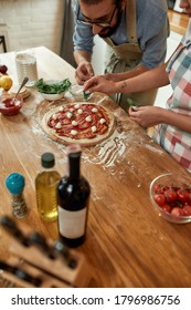 For A Tasty Pizza. Young Couple Making Pizza Together At Home. Man In Apron, Professional Cook Adding Basil On The Dough While Woman Helping Him. Selective Focus On Pizza. High Angle. Vertical Shot