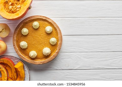 Tasty Pumpkin Pie With Whipped Cream Near Baked And Raw Pumpkins, Whole And Cut Apples On White Wooden Table