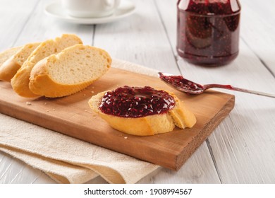 Tasty Sandwich With Raspberry Jam, Baguette Slices And Spoon On A Wood Cutting Board Against Jam Jar Over White Wood Table. Homemade Sweet Red Berry Jelly, Marmalade Or Confiture.