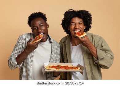 Tasty Snack. Two Hugry Black Guys Eating Italian Pizza From Box And Looking At Camera, Cheerful African American Male Friends Enjoying Food Delivery Service, Standing On Beige Background, Free Space