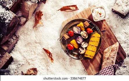 Tasty vegetarian or vegan winter fare with grilled veggie kebabs and corn on the cob on a vintage pewter plate served outdoors in the snow with bread and a savory dip, overhead view