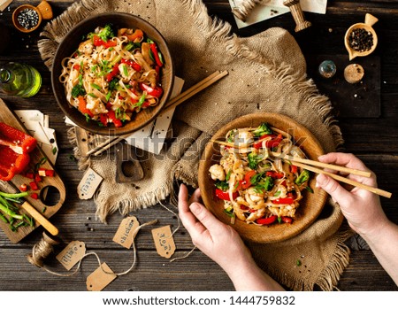 tasty wok noodles with fried chicken fillet, broccoli, red paprika, green onion, sesame in two wooden bowls, woman hands with chopsticks on rustic table with sackcloth, cut veggies, knife, top view