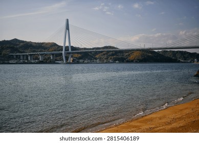 Tatara cable-stayed bridge spanning over blue sea with sandy beach and green hills in background. Coastal landscape on a clear day in Japan.