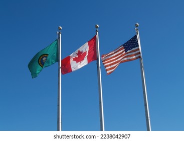 Tattered United States Falg With Canada And Washington State  On Three Flagpoles Against Blue Sky