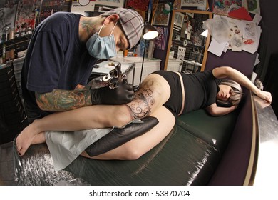 A Tattoo Artist Applying His Craft Onto The Leg Of A Female. Property Release Supplied Includes Tattooists' Studio Hanging Artwork And His Tattoo Work. Shot With Fisheye Lens.