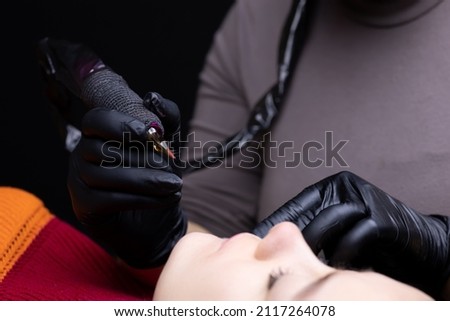 the tattoo artist in sterile gloves holds a tattoo machine in front of the model's face to prepare for the procedure