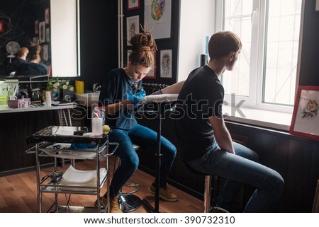 Tattoo girl with dreadlocks on his head in the stuffing sleeve tattoo in black and white. Master works in a professional salon on a white mat and in sterile blue gloves.