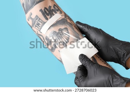 Tattoo master in gloves applying protective film cover over fresh tattoo on man's arm against blue background, closeup