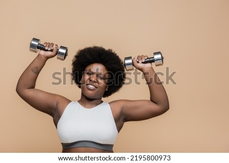 tattooed african american plus size woman in sportswear working out with dumbbells isolated on beige