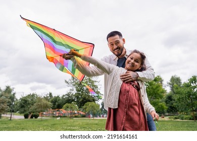 Tattooed Asian Dad Hugging Daughter With Flying Kite And Looking At Camera In Park