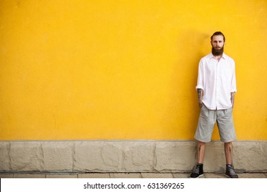 Tattooed Bearded Hipster On Yellow Wall Background Posing Outdoor. Cool Fashion Man In Summer Shorts And White Shirt. Colorful, Trendy And Fashionable Guy