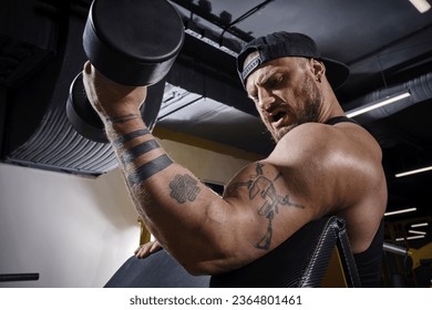 Tattooed, bearded man in black vest and cap. He is lifting a dumbbell, training his biceps, sitting on preacher curl bench at dark gym. Close up - Shutterstock ID 2364801461