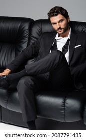 Tattooed And Bearded Man In Formal Wear Sitting On Black Couch Isolated On Grey