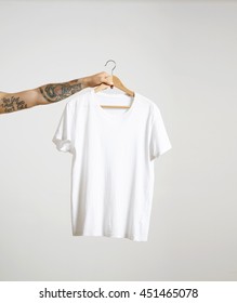 Tattooed biker hand holds hang with blank white t-shirt from premium thin cotton, isolated on white mockup