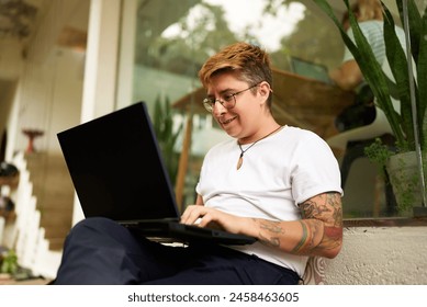 Tattooed Gen Z transgender person works on laptop in cozy office space, displays casual, inclusive workspace. Focused yet comfortable, represents modern work-life balance, LGBTQ-friendly environment. - Powered by Shutterstock