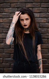 Tattooed girl wearing casual  black clothes, standing against the stone wall or brick fence and playing with her long hair. Brunette young female model with tattoos and piercing in nose