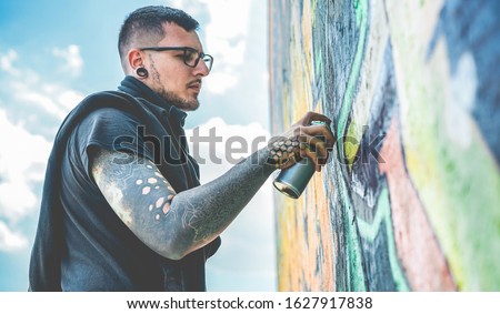 Tattooed graffiti writer painting with color spray his dark picture on the wall - Contemporary artist at work - Urban lifestyle,street art concept