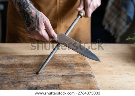 Tattooed hands sharpening a knife on a honing rod over a wooden cutting board, showcasing kitchen preparation skills.