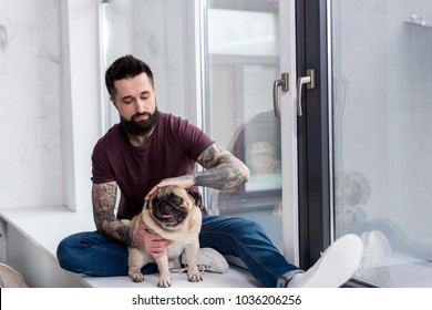 Tattooed Handsome Man Sitting On Windowsill And Palming Dog At Home