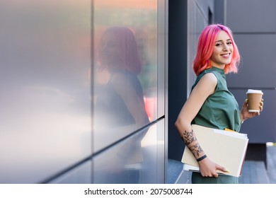 tattooed and happy businesswoman with pink hair holding folder, laptop and coffee to go outside - Powered by Shutterstock