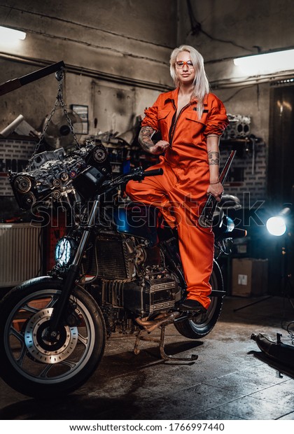 Tattooed hipster girl in\
orange overalls hold a big wrench and posing for a camera while\
standing on her naked bike in garage or workshop, smiling and\
looking on camera