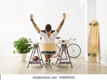 Tattooed lucky freelancer in front of his working space, surrounded with his hobby toys longboard, vintage bicycle and green plant, stretching his hand in air while making break