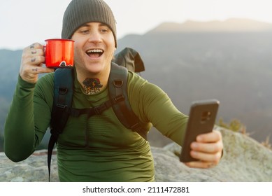 Tattooed Male Hiker With Backpack Takes A Selfie Happily Holding A Red Mug During A Break On His Hike. Pilgrim Doing The Camino De Santiago In A Section Of Asturias, Spain.