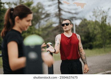 A tattooed man in a red tank top provides guidance to a young woman during an outdoor fitness session in a park, fostering teamwork and fitness engagement. - Powered by Shutterstock