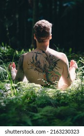 Tattooed Man Sitting On The Grass And Meditating In The Forest. Vertical