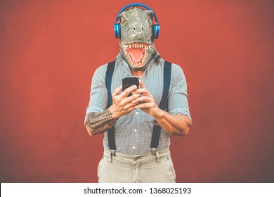 Tattooed man with t-rex mask using smartphone while listening music - Crazy senior guy choosing playlist from mobile phone app - Technology trends and madness costume concept - Focus on face - Powered by Shutterstock