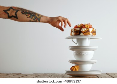 Tattooed Man's Hand Reaching Out To Grab A Piece Of Cake Balanced On A Pyramid Of White Teaware