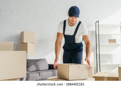 Tattooed Mover In Overalls And Cap Reaching Box In Apartment 