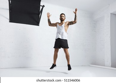 Tattooed And Muscular Athlete Doing Jumping Jacks Isolated On White Brick Wall Next To Black Pull Bar, Looking Left Side