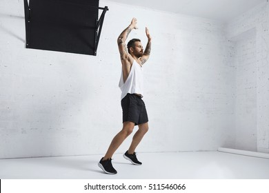 Tattooed And Muscular Athlete Doing Jumping Jacks Isolated On White Brick Wall Next To Black Pull Bar, Looking Right Side